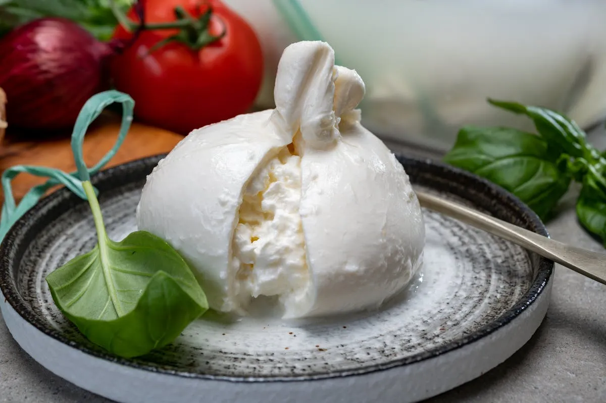 Plan rapproché d'une burrata découpée sur une assiette en céramique, cœur crémeux qui s'écoule, tomates cerises et basilic en arrière-plan